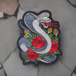 May include: Embroidered patch depicting a cobra with a grey and white body, red tongue, and yellow eyes. The snake is entwined with red roses and green leaves, set against a black border.