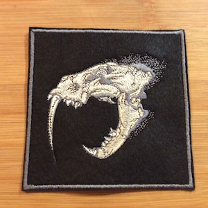 May include: Embroidered patch depicting a detailed saber-toothed tiger skull in grey and cream. The skull is on a black square patch with a grey border, creating a strong contrast. Suitable for application on jackets, bags, or other textiles.