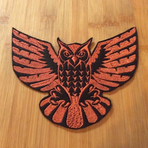 May include: Embroidered patch featuring a detailed owl design with outstretched wings. The owl is rendered in orange thread against a black background, showcasing intricate feather details. A decorative item for clothing or accessories.