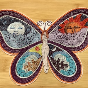 May include: Embroidered butterfly patch with a woman figure at the center. The wings feature celestial and natural elements: a sleeping moon, a sun, and ocean waves. The patch has a purple border with orange accents.