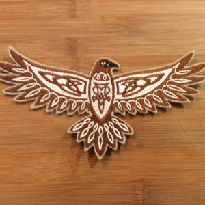May include: Embroidered patch featuring a brown and white stylized eagle with outstretched wings. The eagle has intricate Celtic knotwork designs on its body and wings, with a detailed outline. The patch is likely for clothing or accessories.