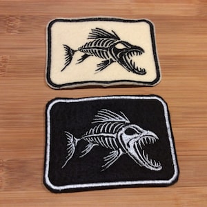 Peut inclure: Deux écussons rectangulaires avec un motif de squelette de poisson. L'un est noir avec un squelette brodé blanc et un bord blanc. L'autre est blanc cassé avec un squelette brodé noir et un bord noir.