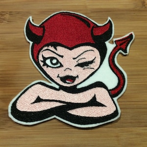 May include: An embroidered patch featuring a cartoon character with pale skin, black hair, and red devil horns. The character has one eye open and one winking, with arms crossed over the chest. A red devil tail extends to the right side.