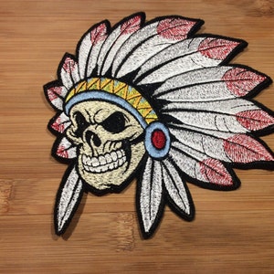 May include: Embroidered patch featuring a skull wearing a Native American headdress. The headdress has white feathers with red tips, a blue and yellow band, and a red and blue circular accent. The skull has a wide grin.