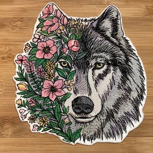 May include: Embroidered patch depicting a wolf's face, with the left side decorated with pink flowers, green leaves, and yellow details. The wolf's fur is in shades of grey and white, with yellow eyes.
