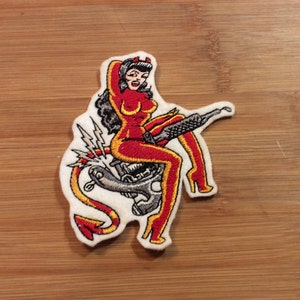 May include: Embroidered patch depicting a red devil woman on a motorcycle. The woman has black hair, red horns, and wears a red outfit with yellow detailing. The motorcycle is grey, and the patch has a white border.