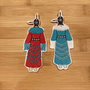 Peut inclure: Deux écussons brodés représentant des personnages stylisés en tenue amérindienne traditionnelle. L'un est rouge et turquoise, l'autre turquoise et blanc. Les deux ont des coutures et des franges détaillées.