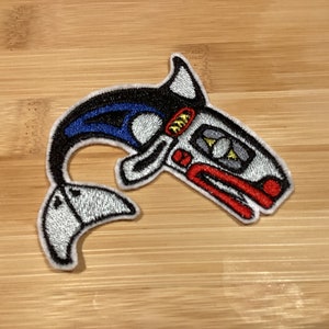 May include: Embroidered patch with a stylized orca whale design. The patch features a black and white body with blue accents, a red mouth, and yellow details. The patch is suitable for clothing or accessories.