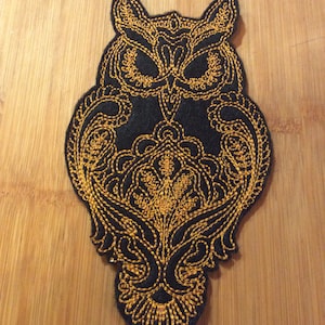 May include: A black owl-shaped patch with intricate gold embroidery. The design features detailed patterns within the owl's body and wings, creating a decorative and ornate appearance. The patch is likely for clothing or accessories.