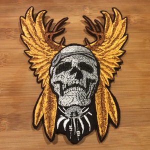 May include: Embroidered patch featuring a skull with a bandana, antlers, and feathered wings in shades of gray, brown, and gold. The patch has a black border and a spider-like design below the skull. The patch is on a wooden surface.