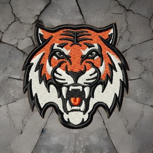 Peut inclure: Écusson brodé représentant une tête de tigre aux couleurs orange, noir et blanc. Le tigre a la gueule ouverte, montrant ses dents et sa langue rouge. Le patch est bordé de noir et a un aspect texturé, idéal pour les vêtements.