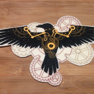 May include: Embroidered patch featuring a black raven with outstretched wings, detailed with gold and cream-colored gears and accents. The raven is set against a backdrop of cream-colored gears with red stitching, creating a steampunk aesthetic.