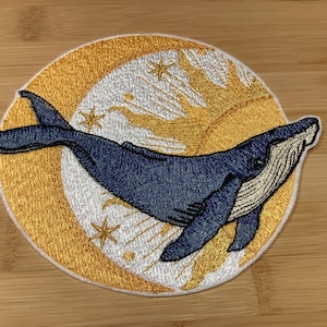 May include: Embroidered patch depicting a blue whale swimming across a gold and white celestial design. The patch has a white border and is likely for clothing or accessories.