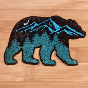 May include: Embroidered bear-shaped patch with a mountain and forest design. The bear is black with teal detailing, including mountains, trees, and a crescent moon. Ideal for clothing or accessories, the patch is likely to be sewn on.