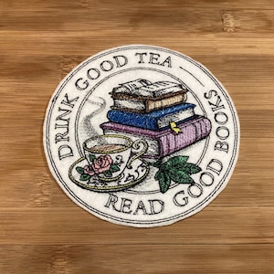 May include: Circular white embroidered coaster with a teacup, books, and the text "DRINK GOOD TEA READ GOOD BOOKS". The teacup has a rose design. The coaster is on a wooden surface. The design is in a circular pattern.