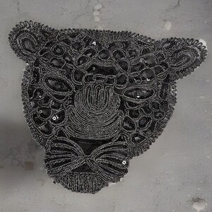 May include: A close-up of a black beaded leopard head. The design features intricate beadwork and sequins, forming the leopard's face. The piece is set against a neutral background, showcasing the detailed craftsmanship and texture of the design.