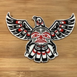 May include: Embroidered patch featuring a stylized eagle design in black, white, red, and gray. The eagle has outstretched wings and a detailed face, showcasing Native American art. The patch is likely for clothing or accessories.