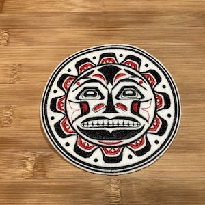 May include: Circular patch featuring a stylized sun face design in black, red, and white. The sun has a smiling mouth, eyes, and radiating lines. The patch is on a wooden surface.