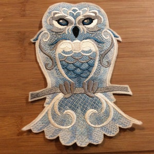May include: An embroidered patch featuring a stylized owl in shades of light blue, white, and grey. The owl has detailed feather patterns, bright blue eyes, and is perched on a horizontal branch. The design includes intricate scrollwork accents.