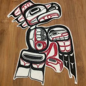 May include: A white, black, and red Native American-style patch featuring a stylized bird design. The patch has a detailed, symmetrical design with black outlines and red accents. The patch is placed on a wooden surface.