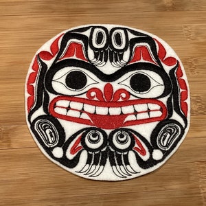 May include: A round, white patch with a Native American-inspired design. The design features a face with black, red, and white colors. The face has large eyes, a wide mouth, and intricate patterns. The patch is on a wooden surface.