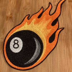 May include: Embroidered patch depicting a black eight ball with the number 8 in white, encircled by orange and yellow flames. The patch has a black border and is designed for application to fabric.