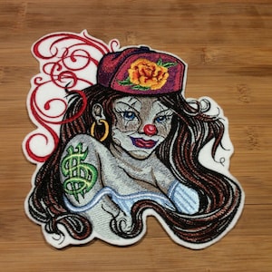 May include: Embroidered patch featuring a female clown with long brown hair, blue eyes, and a red nose. She wears a maroon cap with a yellow rose and a green dollar sign tattoo. Gold hoop earrings and red lipstick complete the design.