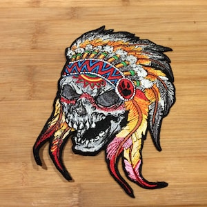 May include: Embroidered patch featuring a skull wearing a Native American headdress. The skull is white with black eye sockets and a wide open mouth. The headdress is adorned with colorful feathers in shades of orange, yellow, and red.