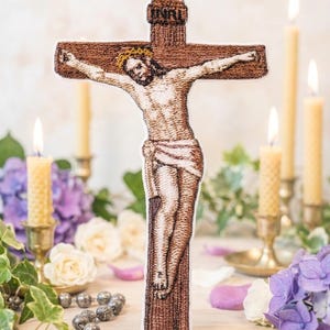May include: Embroidered depiction of Jesus on a cross, with the inscription INRI at the top. The cross is brown, and the figure of Jesus is detailed with a crown of thorns. The background includes lit candles, flowers, and a rosary.
