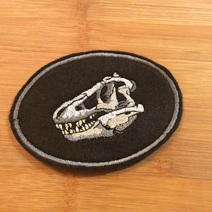 May include: Embroidered patch depicting a Tyrannosaurus Rex skull in grey and yellow on a black oval. The patch has a grey border. Ideal for customising clothing or accessories such as jackets, bags, or other fabric items.