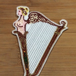 May include: Embroidered patch depicting a stylized figure with a harp. The figure has blonde hair and is partially nude. The harp is white with red and gold detailing. The patch is on a wooden surface.
