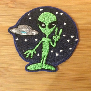 Könnte beinhalten: Gestickter Aufnäher mit einem grünen Alien mit großen schwarzen Augen, das ein Friedenszeichen gibt. Der Alien befindet sich vor einem dunkelblauen Hintergrund mit einer silbernen UFO und weißen Sternen. Der Aufnäher hat einen blauen Rand.