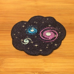 May include: Embroidered patch in the shape of a cloud, featuring a galaxy design. The patch has a dark blue background with embroidered galaxies in blue and purple, surrounded by small gold stars. The patch is on a wooden surface.