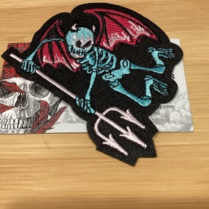 May include: Embroidered patch featuring a turquoise skeleton with red bat wings, holding a pink trident. The patch is on a black background. A card with a skull and roses is partially visible.