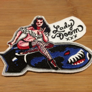 May include: Embroidered patch featuring a devilish woman riding a bomb. The patch has a white border and includes the text "Lady Doom" in black script. The bomb is blue and black with a white shark mouth design.