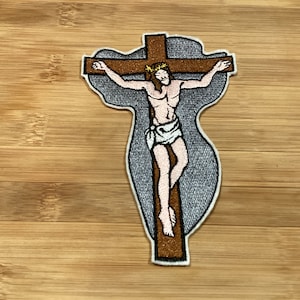 May include: Embroidered patch depicting Jesus on the cross. The figure is brown with a white loincloth and a crown of thorns. The cross is brown with a gray outline, set against a gray background.