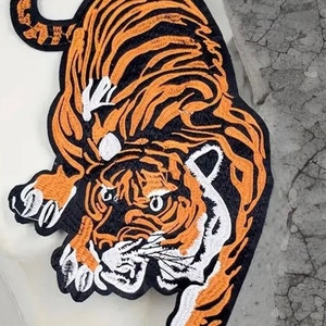 May include: Embroidered tiger patch featuring orange and black stripes, white details, and a black border. The tiger is shown in a dynamic pose, with its claws extended. Suitable for application on jackets, bags, or other fabric items.