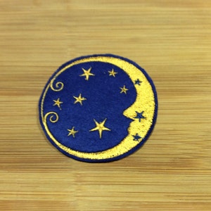 May include: A circular embroidered patch with a crescent moon and stars design. The moon is gold, with a face, and the background is dark blue. Gold stars are scattered around the moon. Suitable for clothing or accessories.