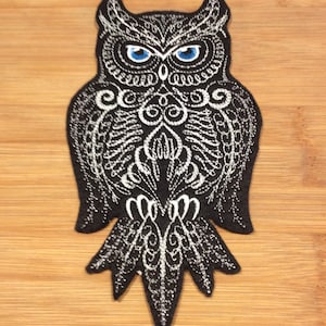 May include: Black owl patch with intricate white embroidered details. The owl has blue eyes and a detailed, symmetrical design. The patch is likely for clothing or accessories, adding a decorative element.