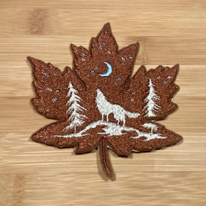 May include: A brown embroidered maple leaf patch. The design features a howling wolf, trees, a crescent moon, and stars. The patch is suitable for clothing or bags. The patch is approximately 8 cm in length.