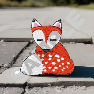 May include: Embroidered patch featuring a stylized fox with closed eyes. The fox has a red body with white and black accents, and a white tail with white spots. The patch is likely for clothing or accessories.