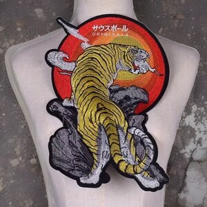 Peut inclure: Écusson brodé représentant un tigre jaune et noir sur une formation rocheuse grise, sur fond circulaire rouge et orange. L'écusson comprend du texte japonais et le mot "ORIGINALS".