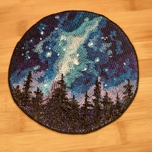 May include: Circular patch featuring a detailed embroidered night sky scene. The design includes a vibrant aurora borealis in shades of blue and green, a starry sky, and a silhouetted forest at the bottom. The patch has a black border.