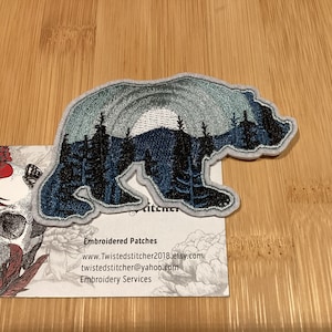 Peut inclure: Écusson brodé en forme d'ours, représentant un paysage de montagne et de forêt dans des tons de bleu et de gris. L'écusson a une bordure blanche. Une carte de visite en dessous présente un crâne et un motif floral.