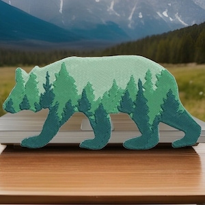 May include: A green bear-shaped patch with a forest design. The bear is walking, with a light green body and a darker green forest silhouette inside. The patch is on a wooden surface with a blurred background of mountains and trees.