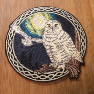 May include: A circular embroidered patch featuring a white snowy owl perched on a brown tree branch against a dark blue night sky with a yellow moon. A second owl is in flight. The design is enclosed by a white and black Celtic knot border.