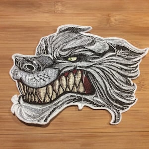 May include: An embroidered patch featuring the head of a snarling wolf in shades of silver, grey, and black. The wolf has bared, sharp, off-white teeth, a dark red mouth interior, and a single visible green eye, all outlined in white thread.