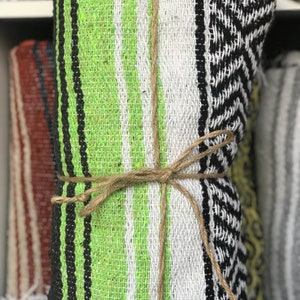 May include: Rolled-up woven blanket with a green, black, and white striped pattern, tied with twine. The blanket has a textured appearance and is part of a collection of similar blankets.