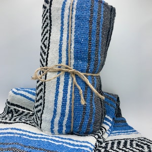 May include: A woven blanket featuring a striped pattern in shades of blue, white, black, and gray. The blanket is folded and tied with a piece of natural jute twine. The fabric displays a textured weave with geometric diamond motifs on the ends.