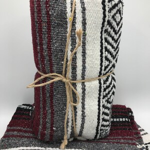 May include: A folded woven blanket with a striped pattern in burgundy, gray, white, and black. The blanket is tied with twine. Another blanket with a similar pattern is beneath the folded one.